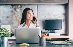 A woman in white shirt talking on phone at her desk with laptop and office supplies.